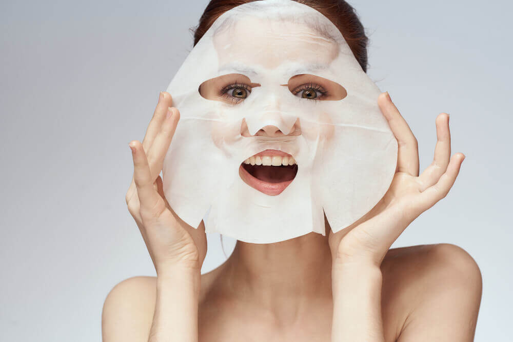 woman smiling and holding a sheet mask in front of her face